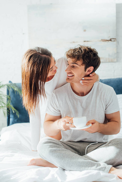 Young adult sitting on bed with cup of coffee and looking at girlfriend - Foto, Imagem