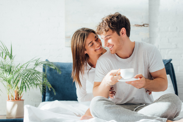 Young woman embracing and looking at boyfriend with cup of coffee on bed - Фото, зображення