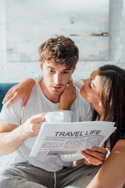 Selective focus of woman embracing boyfriend with cup of coffee and newspaper on bed  - Photo, Image