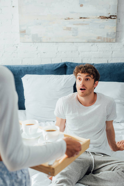 Szelektív fókusz izgatott férfi nézi barátnő csésze kávé reggeli tálcán a hálószobában  - Fotó, kép
