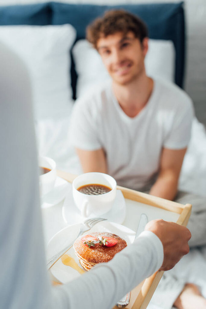 Szelektív fókusz a nő kezében reggeli tálca kávé és palacsinta közelében barátja a hálószobában  - Fotó, kép