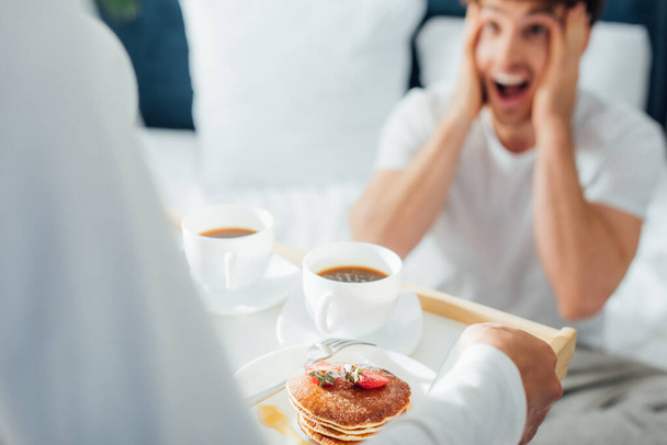 Selective focus of woman holding breakfast and coffee on tray near excited boyfriend in bedroom  - Fotoğraf, Görsel