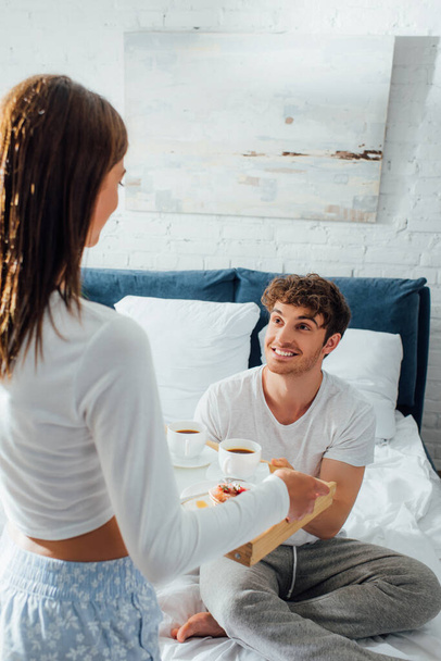 Selective focus of young man looking at girlfriend with breakfast tray in bedroom at morning  - Фото, изображение