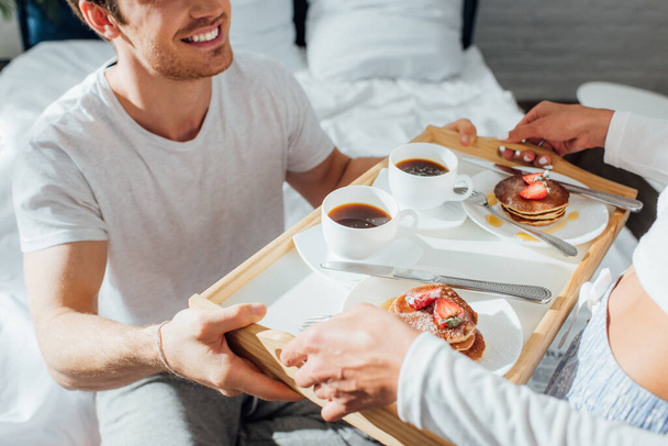 Vista cortada de jovem casal em pijama segurando bandeja de café da manhã com panquecas e café na cama  - Foto, Imagem
