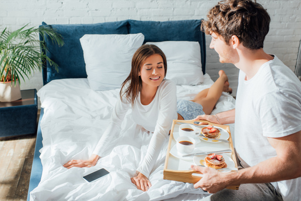 Selective focus of woman in pajamas looking at breakfast on tray near boyfriend in bedroom  - Fotoğraf, Görsel