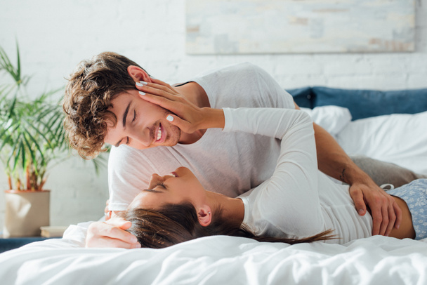 Genç bir kadın erkek arkadaşının yüzüne dokunuyor pijamalarıyla yatakta yatarken.  - Fotoğraf, Görsel