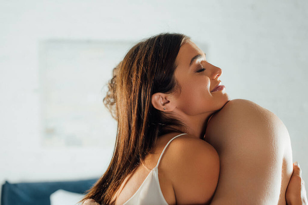 Mujer joven con los ojos cerrados abrazando hombre sin camisa en casa  - Foto, imagen