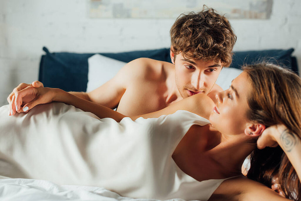 Muscular man kissing shoulder of young girlfriend in silk nightdress on bed  - Fotografie, Obrázek
