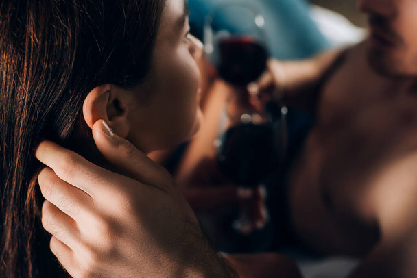 Selektivní zaměření muže bez trička dotýkající se vlasů přítelkyně a držící sklenici vína  - Fotografie, Obrázek