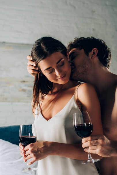 Selective focus of shirtless man kissing seductive woman with glass of wine in bedroom  - Fotoğraf, Görsel