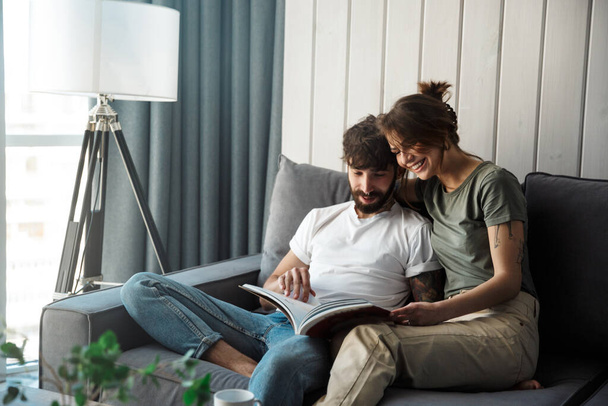 素敵な若いカップルが自宅でソファでリラックスし、雑誌を読む - 写真・画像