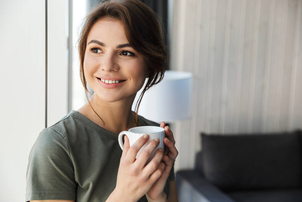 Attractive smiling young woman holding coffee cup while standing at the window indoors - Fotoğraf, Görsel