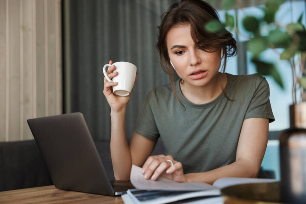 Attractive pensive young woman working on laptop computer while sitting at the table at home, drinking coffee - Photo, Image