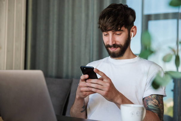 Handsome smiling young bearded man working on laptop computer while sitting at the kitchen table, drinking coffee at home, using mobile phone - 写真・画像