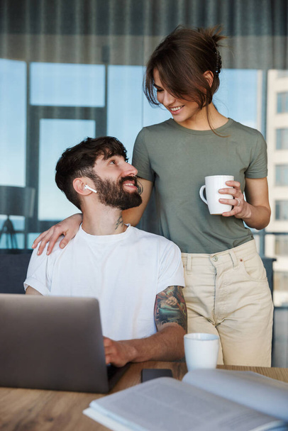 Beautiful smiling young couple using laptop computer while spending time in the kitchen, drinking coffee - Photo, image