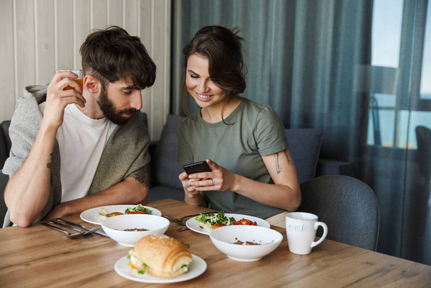 Lovely young couple having healthy breakfast in the kitchen, using mobile phone - Foto, imagen