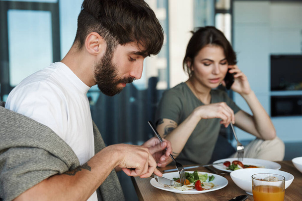 Lovely young couple having healthy breakfast in the kitchen, talking on mobile phone - Photo, Image