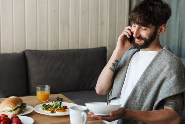 Attractive smiling young bearded man using mobile phone while having healthy tasty dinner at home - Foto, imagen
