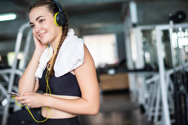 Młoda atrakcyjna kobieta słuchająca muzyki przez słuchawki łączy się ze smartfonem lub telefonem komórkowym. Relaks po ciężkim treningu na siłowni   - Zdjęcie, obraz