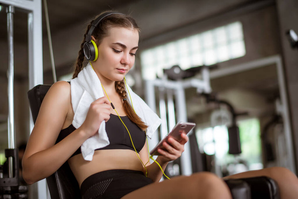 Junge attraktive Frau, die per Kopfhörer Musik hört, verbindet sich mit Smartphone oder Handy. Entspannung nach hartem Training im Fitnessstudio   - Foto, Bild
