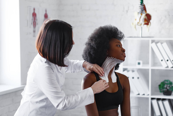 Kinesiology taping. Physiotherapist applying kinesiology tape to patient neck. Therapist treating young female African American athlete. Post traumatic rehabilitation, sport physical therapy - Foto, afbeelding
