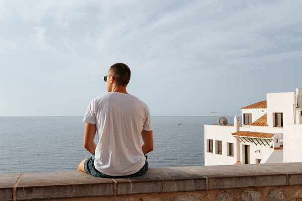 Young man resting looking at the calm sea.Young man enjoying the sea side view.Summer lifestyle image of stunning man sitting above the sea. - 写真・画像
