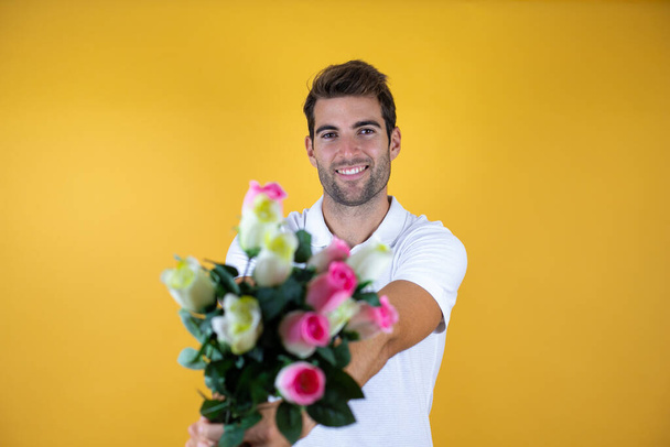 junger gutaussehender Mann, der über gelbem, isoliertem Hintergrund steht und lächelnd Blumen hält - Foto, Bild