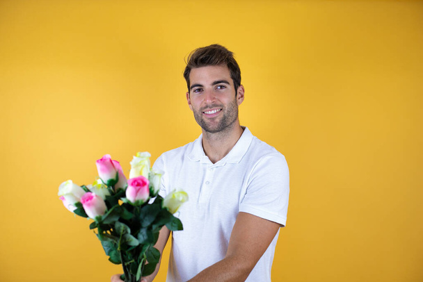 joven guapo hombre de pie sobre amarillo insolado fondo sonriendo sosteniendo flores - Foto, Imagen