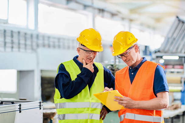 Ein Ingenieur und ein Manager arbeiten in einer Fabrik zusammen und tragen Schutzkleidung - Foto, Bild