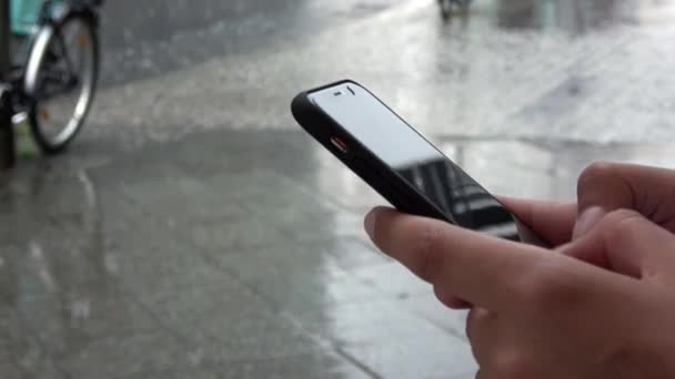 4K. Egy üzletasszony használja az okostelefonját, miközben esik. Üzenetek vagy e-mail küldése. Egy közeli kép, amint gépel a telefonon. Az esőcseppek a földre hullanak. - Felvétel, videó