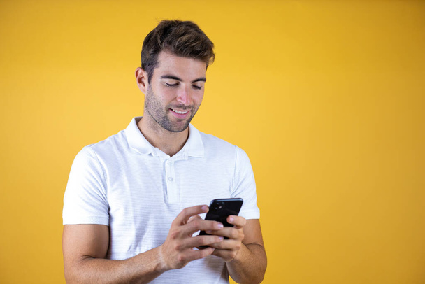 Sarı arka plan üzerinde akıllı telefon kullanan genç bir adam mutlu bir yüz takınıyor ve gülümsüyor. Dişlerini gösteren bir gülümsemeyle. - Fotoğraf, Görsel