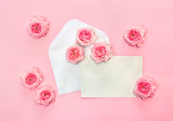 Hermosas flores rosas rosadas en sobre postal y hoja en blanco con espacio para el texto sobre un fondo de papel rosa. Vista superior, plano - Foto, Imagen