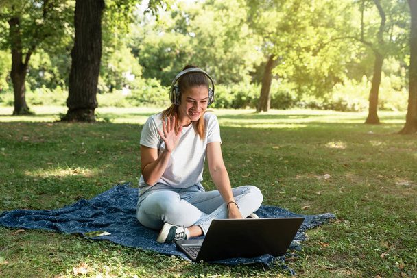 Młoda studentka siedzi w parku ze swoim laptopem i bezprzewodowymi słuchawkami, rozmawiając przez internet z rodzicami i rodziną   - Zdjęcie, obraz