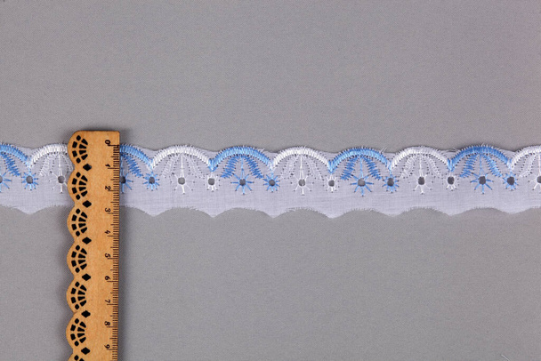 グレーの背景に刺繍や木製の定規と白い青い綿のリボンのストレートストリップ。縫製のための全国的なパターン。パターンと織り糸。ウェブサイトのテクスチャ。テキストのスペース. - 写真・画像