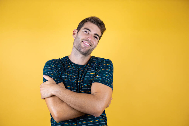 Junger gutaussehender Mann, der vor isoliertem gelben Hintergrund steht und sich glücklich und positiv umarmt, selbstbewusst lächelt - Foto, Bild