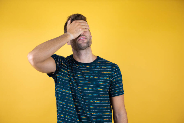 Young handsome man covering face with one hand on yellow background. Wearing a blue t-shirt - Photo, Image