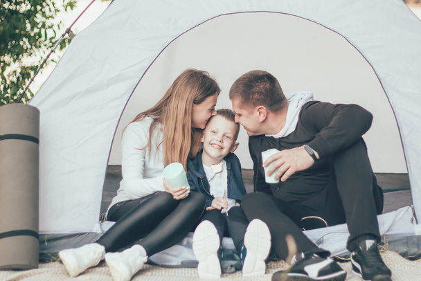 Family sitting in nature near the tent mom and dad kiss their son - Photo, Image