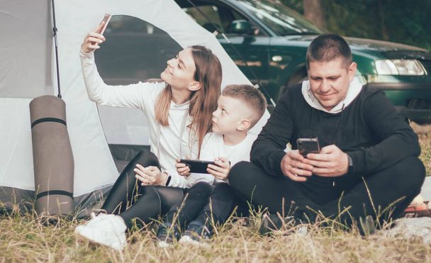  Familia sentada en la naturaleza cerca de la tienda mamá se toma una selfie con su hijo y papá se sienta en el teléfono - Foto, Imagen