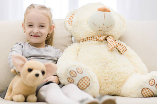 High angle portrait of a young smiling girl with stuffed toy sitting on sofa in the living room at home. - Valokuva, kuva