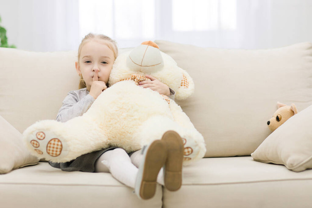 Cute little girl sitting with her legs curled up, teddy bear on her knees. - Foto, afbeelding
