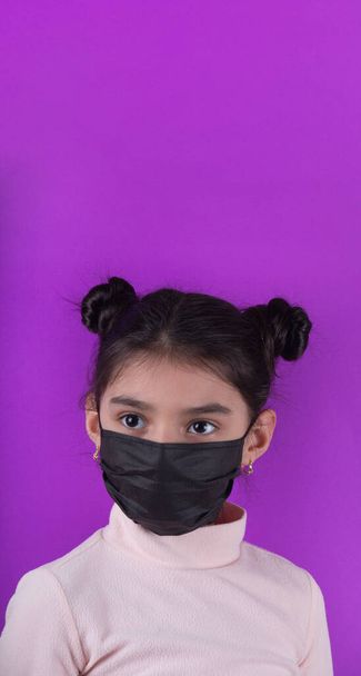 紫の背景に聴診器と黒のマスクを持つかわいい女の子。家にいて。COVY-19を停止します。使い捨てマスク。ウイルス対策装置。隔離だ。女の子は医者を演じる。教育と職業概念です。黒いマスク - 写真・画像