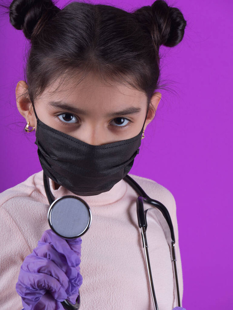 紫色の背景に聴診器とカラーマスクを持つかわいい女の子。家にいて。COVY-19を停止します。ウイルス対策装置。隔離だ。女の子は医者を演じる。教育と職業概念です。じっとしてろ。面白い表現.  - 写真・画像