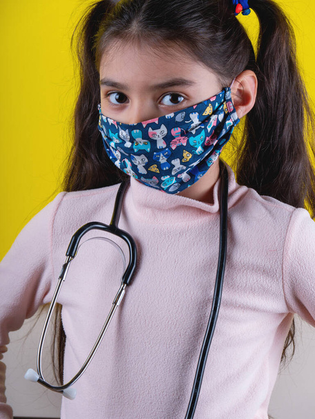 黄色の背景に聴診器とカラーマスクを持つかわいい女の子。家にいて。COVY-19を停止します。ウイルス対策装置。隔離だ。女の子は医者を演じる。教育と職業概念です。じっとして - 写真・画像