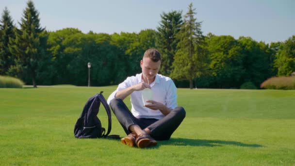 El hombre se sienta en el parque sobre hierba verde, come ensalada vegetariana y la escupe. Chico en camisa blanca, pantalones y mochila sostiene en las manos un paquete de papel ambiental con alimentos y palos de madera - Imágenes, Vídeo