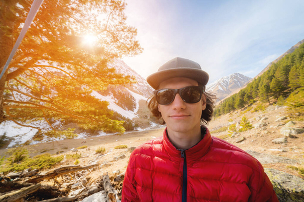 Portrait of a long-haired young man in a red down jacket and a gray cap in sunglasses looks into the camera against the background of a mountain gorge with snow-capped peaks in the distance - Photo, image