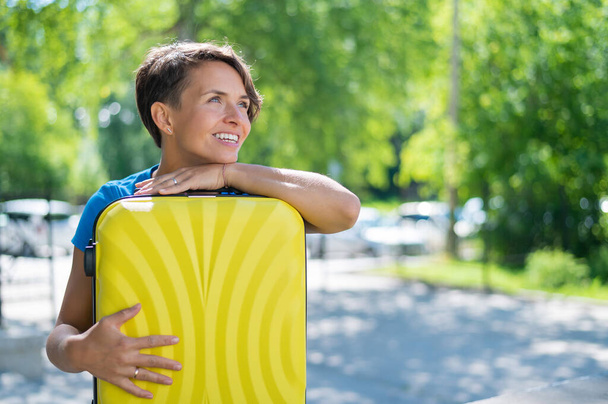 Close-up portrait of a beautiful short-haired Caucasian young woman with a yellow suitcase outdoors. The girl with a perfect smile dreamily leaned her head against a large luggage bag. - Foto, immagini