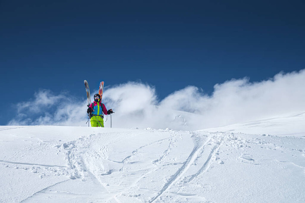 Portrait of a women skier in a multi-colored bright jacket in a ski mask with her face closed on a sunny day against the backdrop of snow-capped Caucasian mountains and clouds - Foto, Imagem