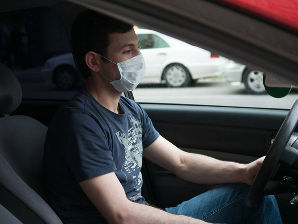 Portrait of man wearing disposable medical facemask in a car during coronavirus outbreak.Young man in protective sterile medical mask driving car.  - Photo, Image