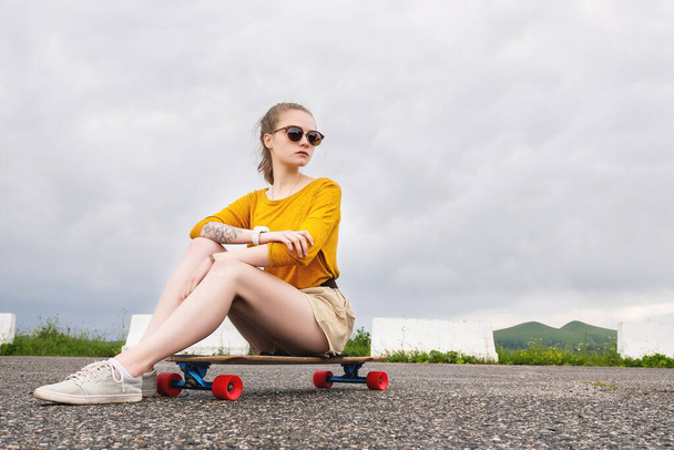 A young attractive girl in a yellow sweater shorts and sunglasses with a tattoo on her arm sits on a longboard behind a suburban asphalt pad - Photo, image