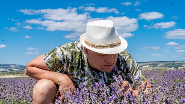 Smiling guy in lilac field having a joyful face sitting in a cap against the blue sky - Фото, изображение